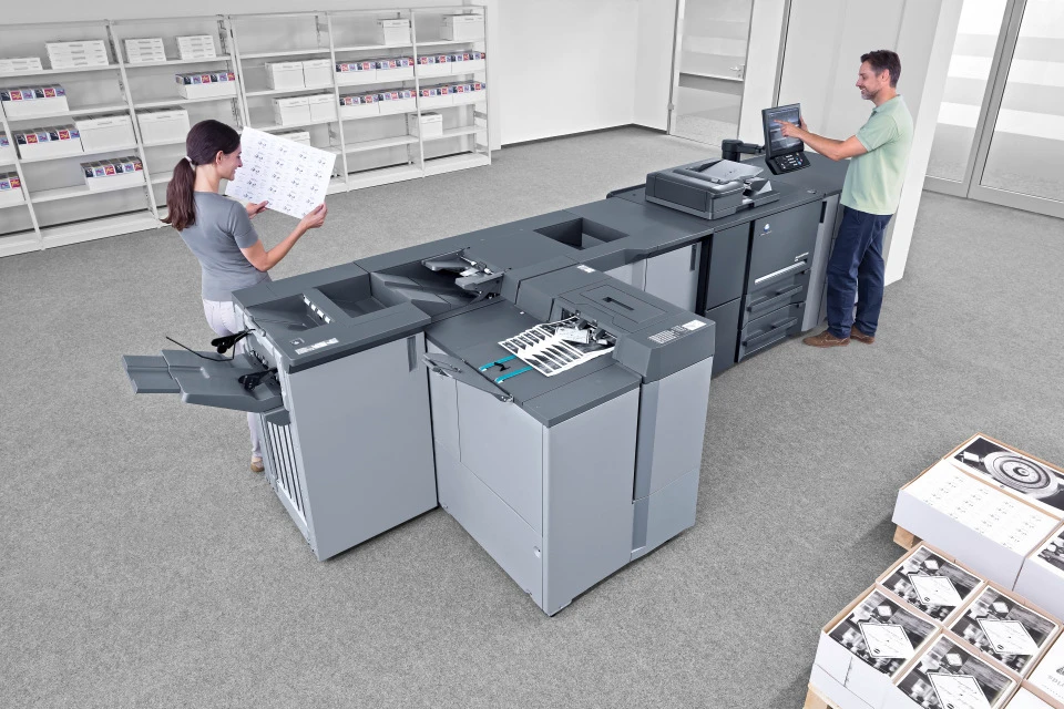 Konica Minolta AccurioPress 6136 Schwarzweiß-Produktionsdrucker mit integrierter Weiterverarbeitung – Mitarbeitende entnehmen und kontrollieren Druckerzeugnisse in einer Hausdruckerei