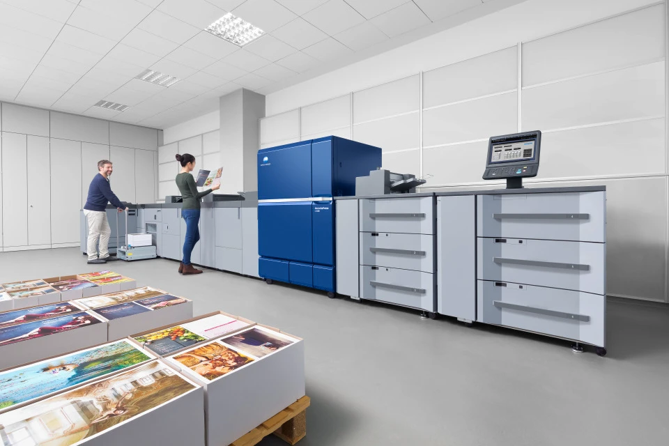 Konica Minolta AccurioPress C12000 Farbproduktionsdrucker in einer Hausdruckerei – zwei Mitarbeitende prüfen hochwertige Farbdrucke auf Paletten
