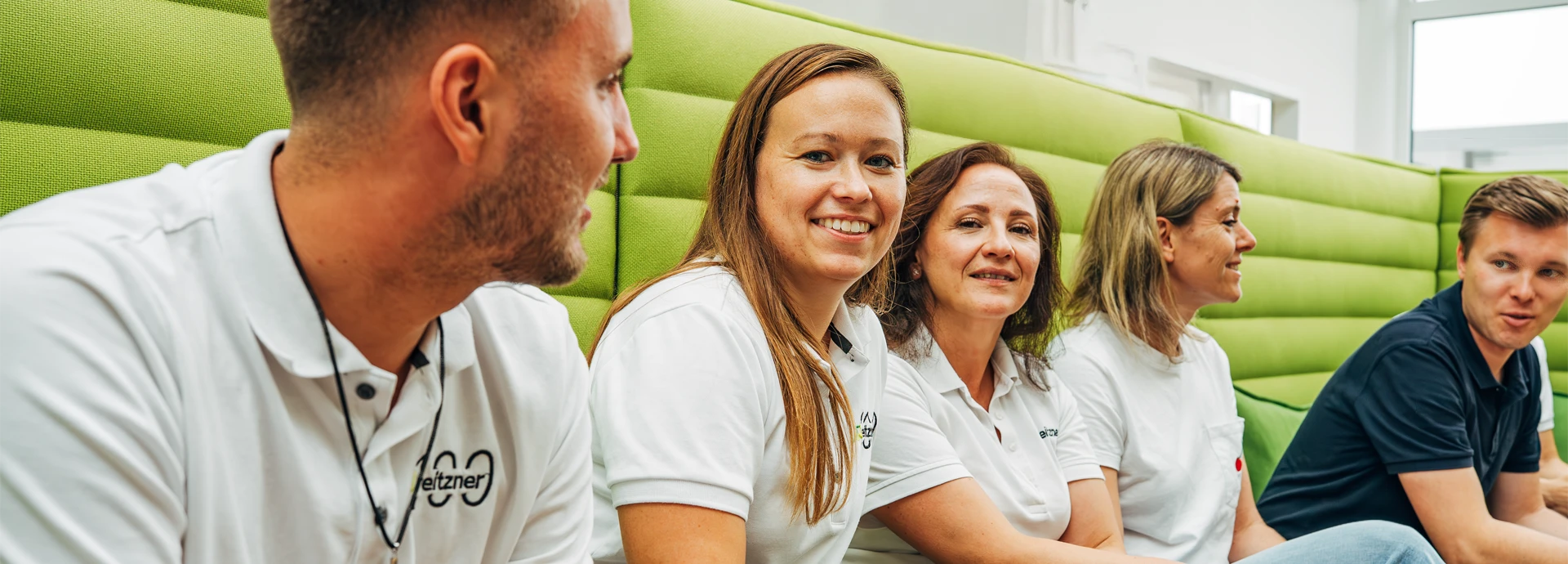 Ein lächelndes Team sitzt in einer modernen Lounge-Ecke und tauscht sich aus. Die offene Kommunikation und flache Hierarchien spiegeln die New Work Kultur im Unternehmen wider.