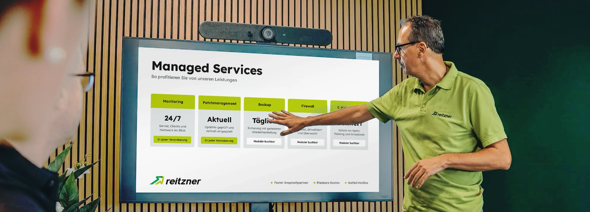 Home 24 Ein reitzner Mitarbeiter zeigt in einer Besprechung auf einen Bildschirm mit den Managed-Services-Bausteinen Monitoring, Backup, E-Mail Security und Firewall
