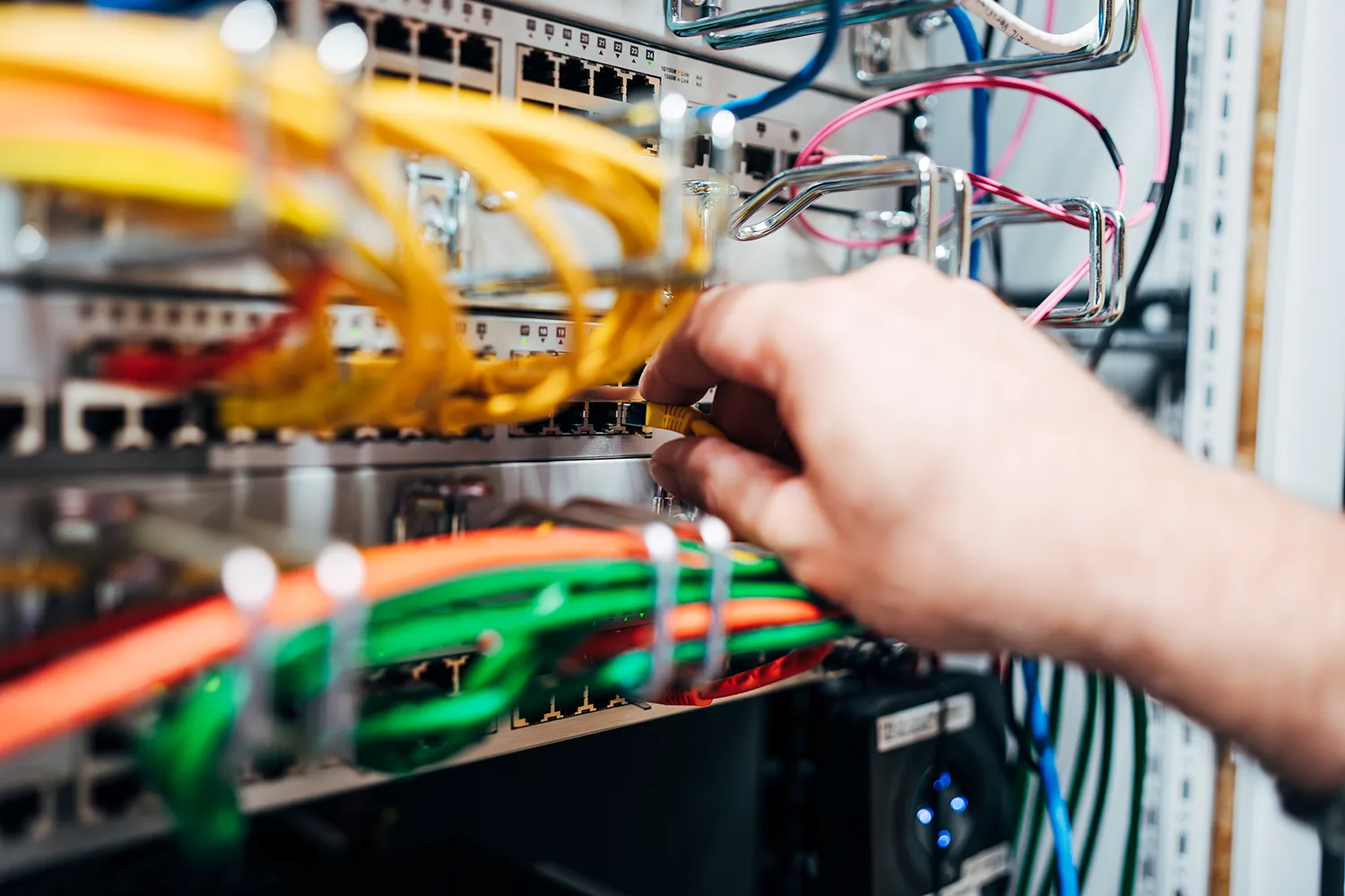 Managed Services: Fokus auf Ihr Kerngeschäft 1 Hand steckt ein gelbes Netzwerkkabel in einen Switch im Serverschrank, daneben farbig sortierte Patchkabel