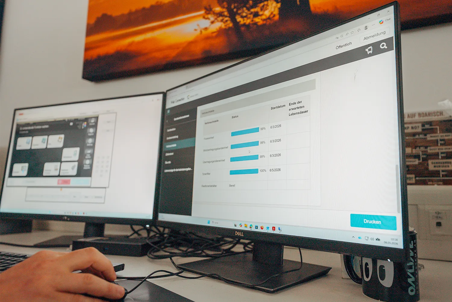 Zwei Monitore am Arbeitsplatz zeigen das Statusdashboard einer Geräteüberwachung mit Füllständen und Restlaufzeiten, daneben eine Hand an der Maus